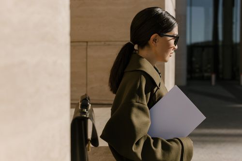 Free Side view of a woman in an olive coat holding a document, standing outdoors. Stock Photo