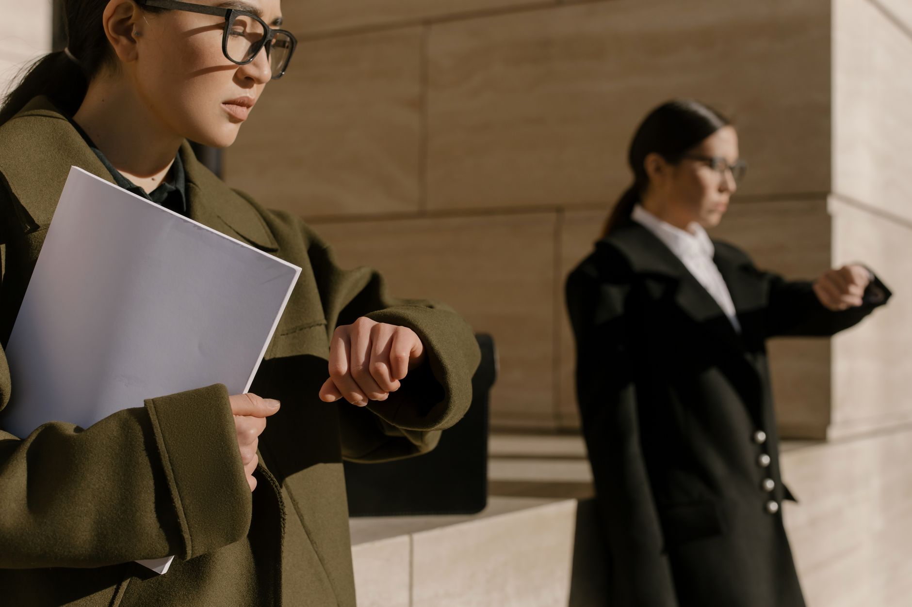 Two businesswomen wearing coats, checking time in bright daylight.