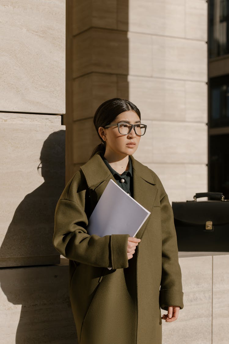 Pretty Woman Holding A White Folder Standing Near A Wall