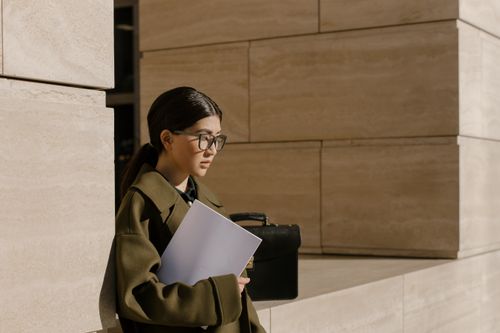 Free An Asian woman in eyeglasses holding a folder in an urban outdoor setting, exuding professionalism. Stock Photo