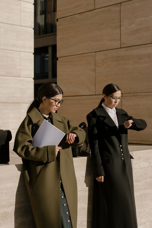 Free Two fashionable women in long coats checking time under sunlight outdoors. Stock Photo