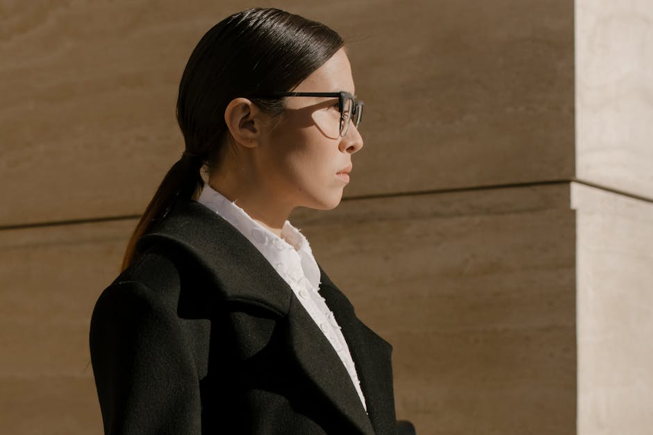 A stylish woman in sunglasses with sleek hair stands outdoors in modern attire.