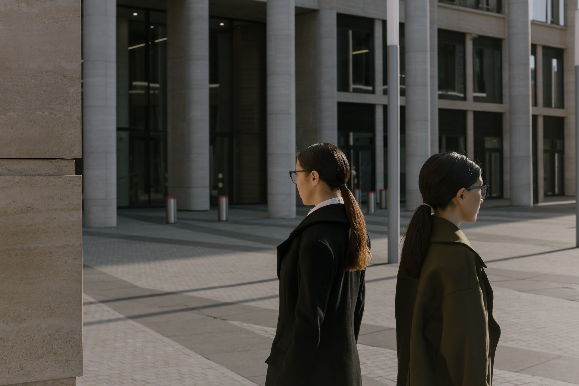 Two professional women in coats walking near a modern urban office building during the day.