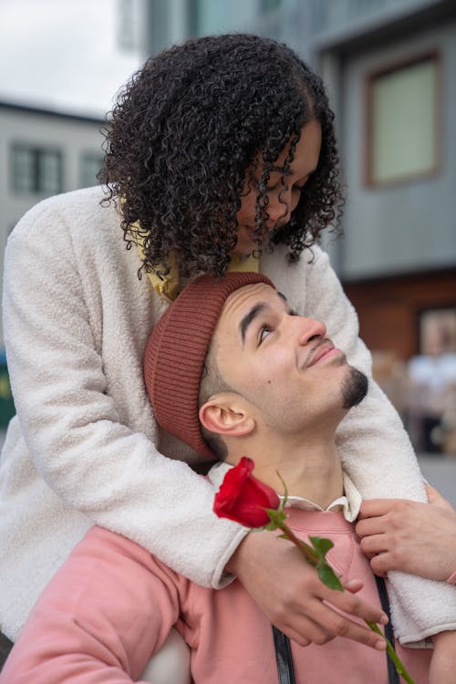 Loving ethnic female with curly hair holding tender red rose standing behind and embracing content young boyfriend on street