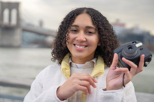 Free Smiling young Hispanic female in warm jacket standing with photo camera on city street and looking at camera on cloudy overcast day Stock Photo