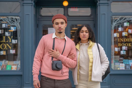 Stylish young ethnic female and male travelers in stylish outfits standing together on city street near aged bookstore and looking at camera