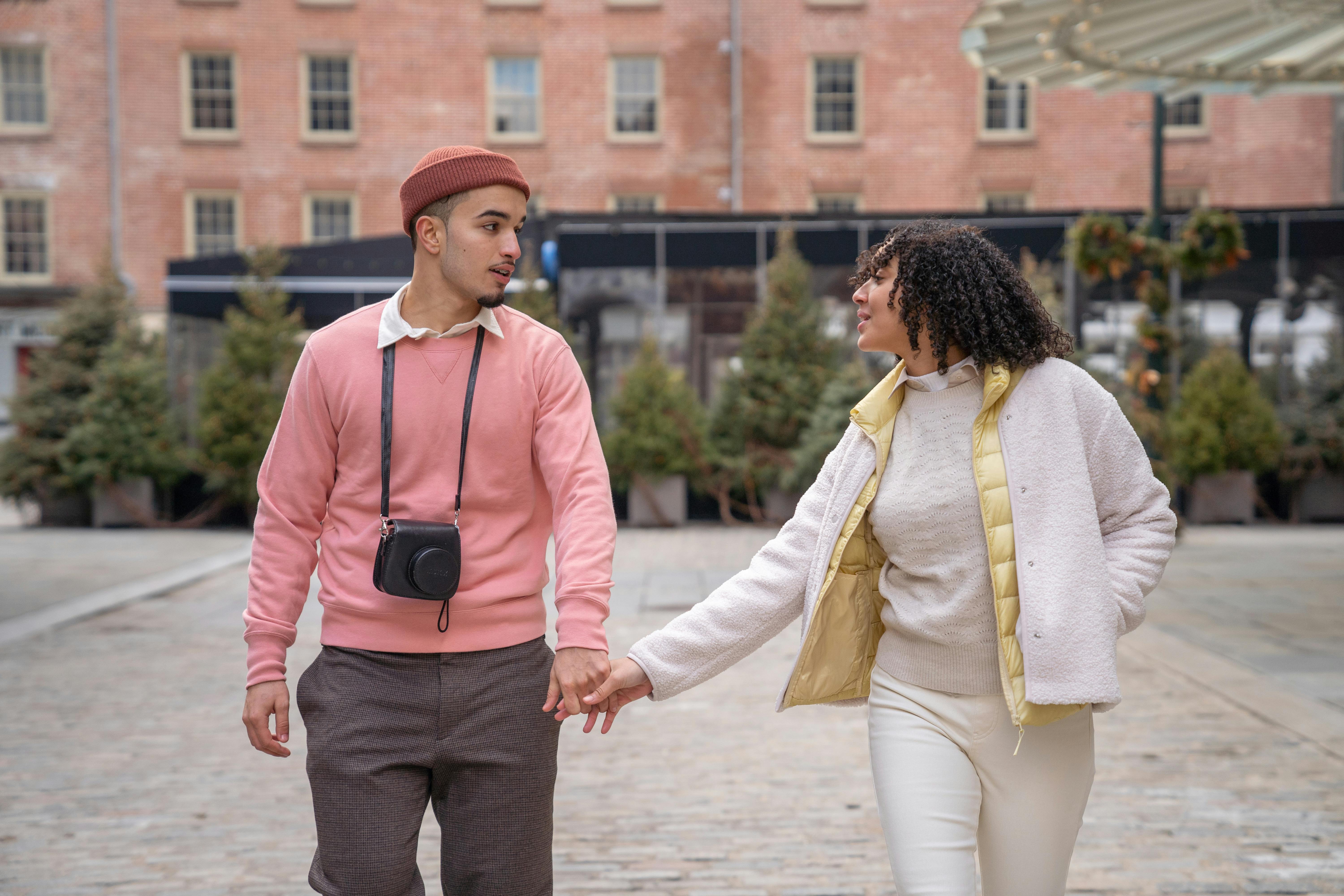Positive young ethnic couple in stylish warm clothes holding hands while walking in aged district of city during vacation and looking at each other