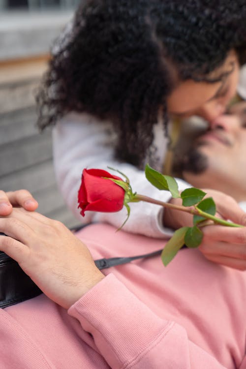 Free From above of young ethnic female millennial with fresh red rose in hand hugging and kissing happy boyfriend during romantic date in park Stock Photo