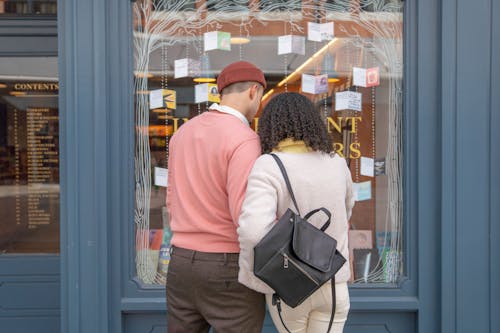 Back view of unrecognizable young couple in stylish outfits choosing books while standing on street near glass showcase of store during weekend