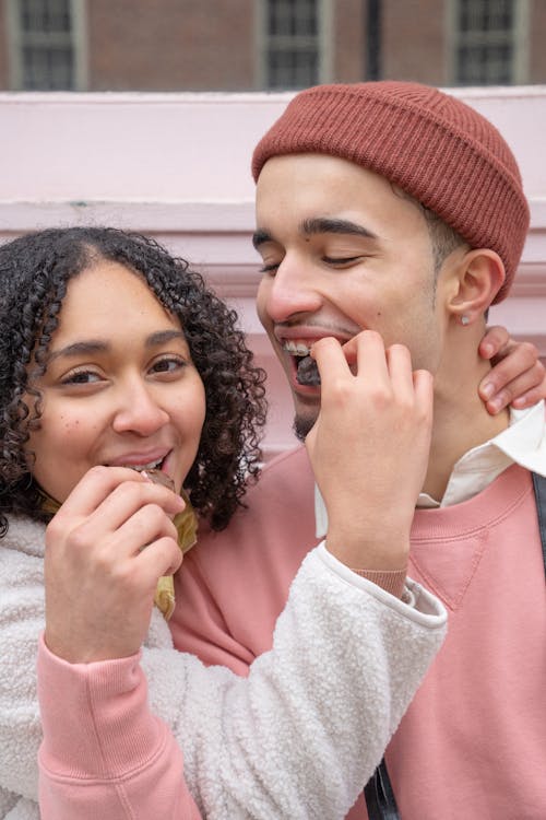 Free Cheerful young Hispanic couple in casual outfits smiling and feeding each other with yummy chocolates while spending time together on city street Stock Photo