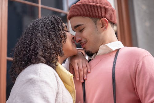 Free Side view of loving young Hispanic couple in stylish clothes touching noses with closed eyes and smiling while standing on city street on sunny day Stock Photo