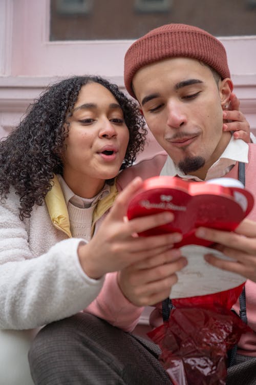 Free Positive Hispanic couple with heart shaped box of delicious candies sitting on street on city while celebrating Saint Valentines day Stock Photo