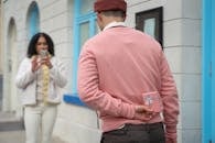 Blurred Hispanic woman taking photo of faceless boyfriend with pink gift box behind back during romantic date on street near building