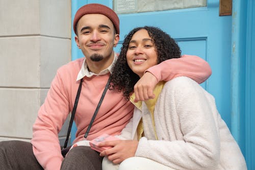 Smiling Hispanic boyfriend hugging girlfriend with gift box while sitting on street near building and looking at camera during date