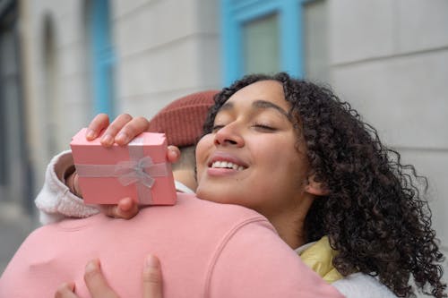 Content Hispanic girlfriend with gift box in hand embracing anonymous boyfriend while standing on street on blurred background during romantic date