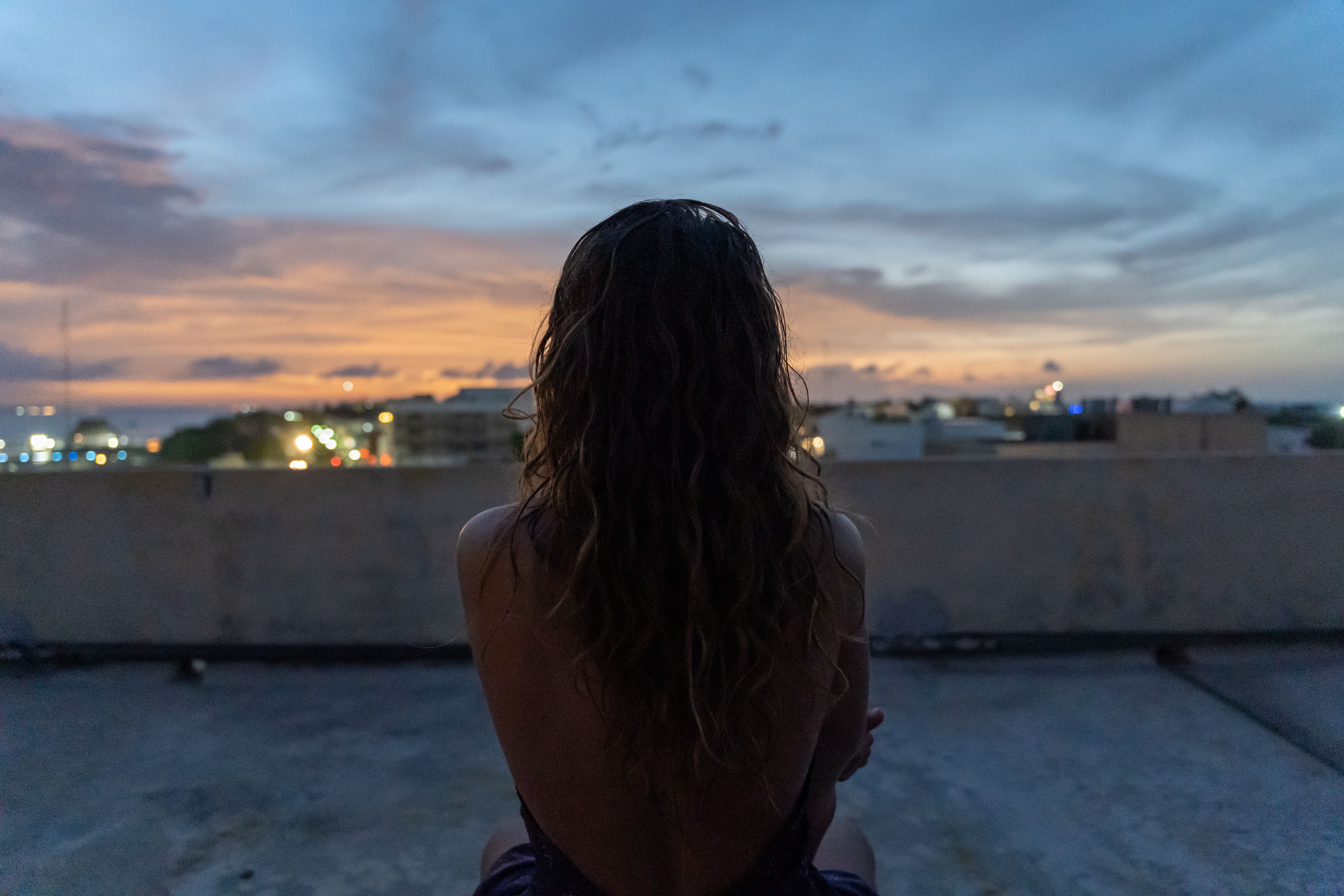Back view of a woman with curly hair admiring a vibrant city sunset from a rooftop.