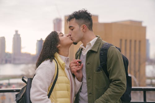 Side view of romantic Hispanic couple with backpacks in casual wear kissing while caressing on street with buildings during date