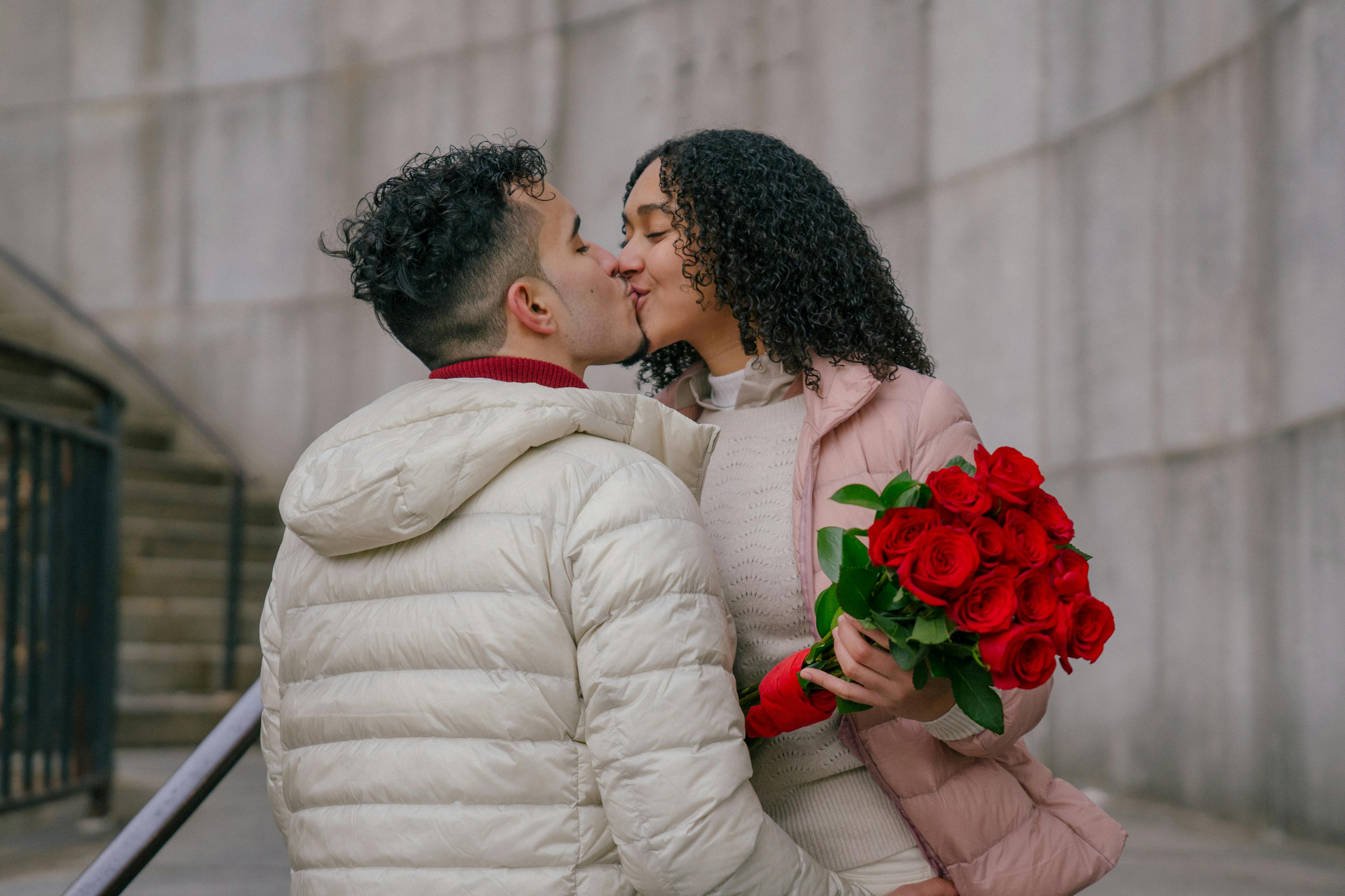 Loving ethnic couple with bouquet of bright blooming roses kissing each other on urban street