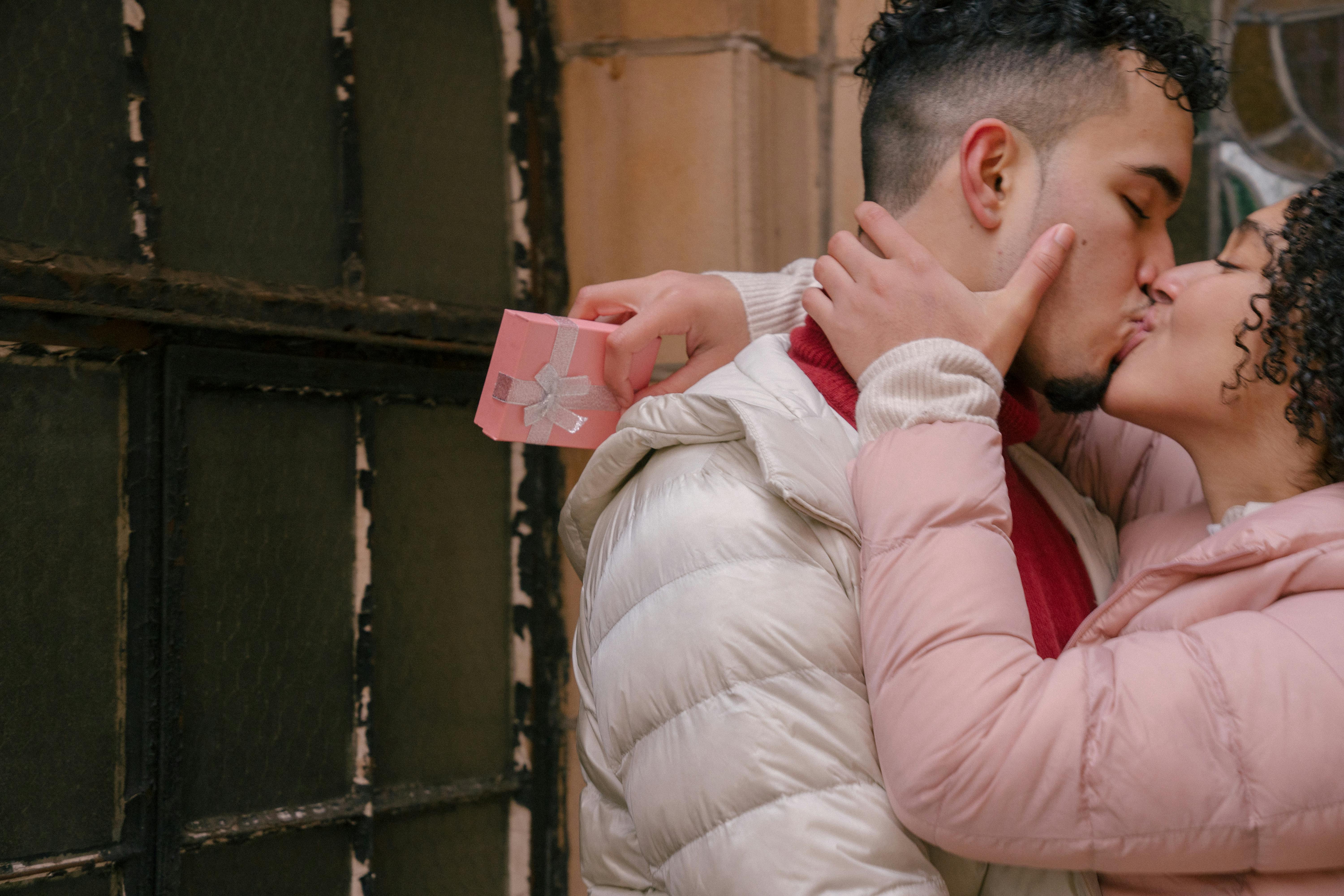 Side view of crop ethnic couple with festive box of present kissing and embracing near fence