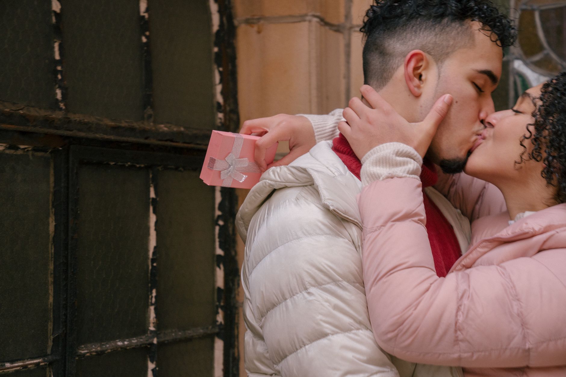 Side view of crop ethnic couple with festive box of present kissing and embracing near fence