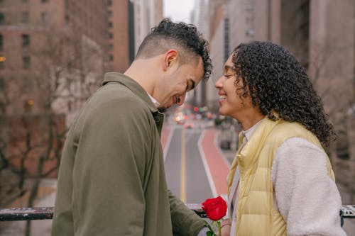 Side view of cheerful ethnic couple with bright red blooming flower smiling on blurred background of urban street