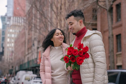 Free Young glad ethnic couple with bouquet of bright red roses smiling on urban street Stock Photo