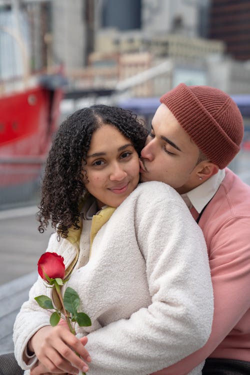 Free Young ethnic boyfriend kissing cheerful girlfriend with bright red blooming rose on blurred background of town Stock Photo