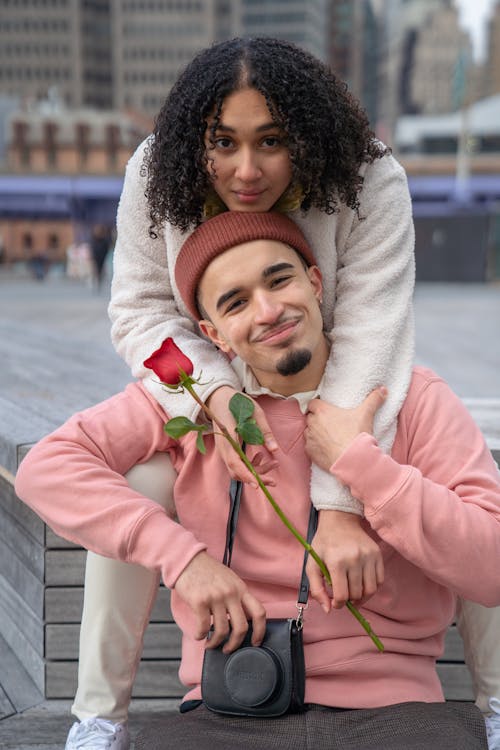 Free Cheerful young ethnic couple with red rose in blossom hugging and looking at camera on blurred background of town Stock Photo