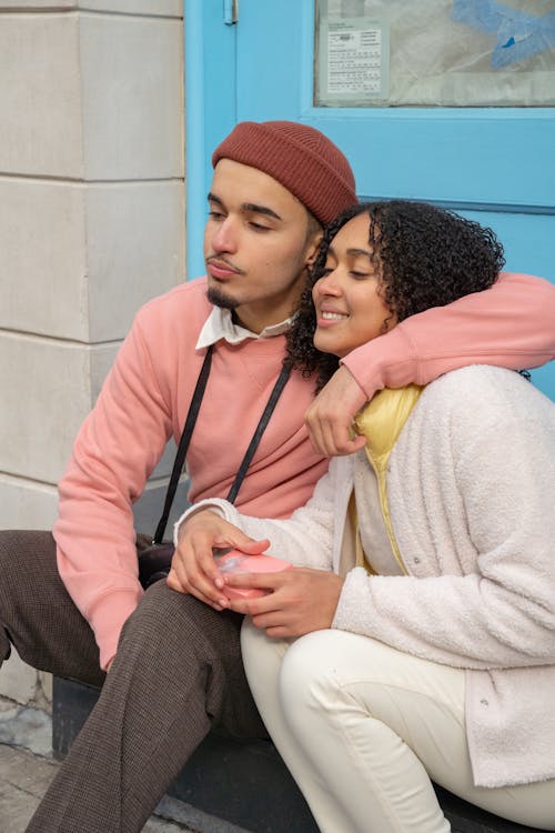 Free Confident young ethnic male hugging smiling girlfriend while sitting together on step of aged building on street Stock Photo