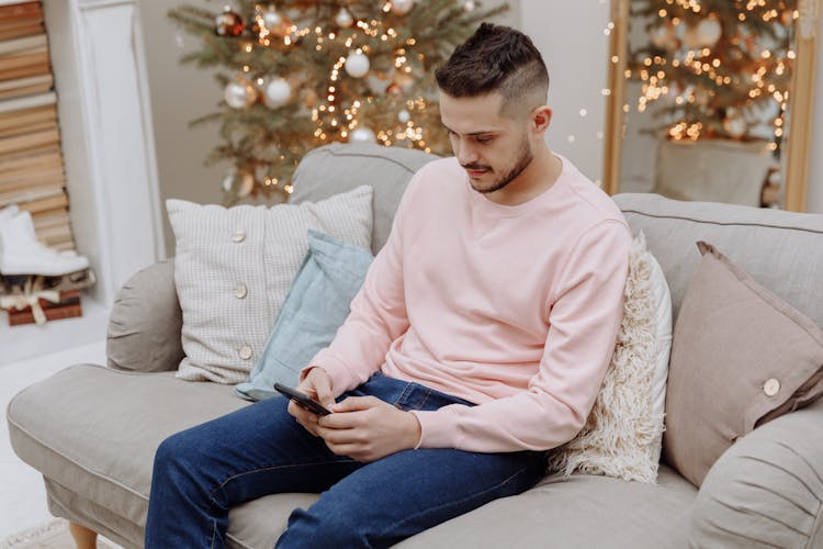 A Man In Pink Sweater And Denim Jeans Sitting On The Couch While Using His Mobile Phone