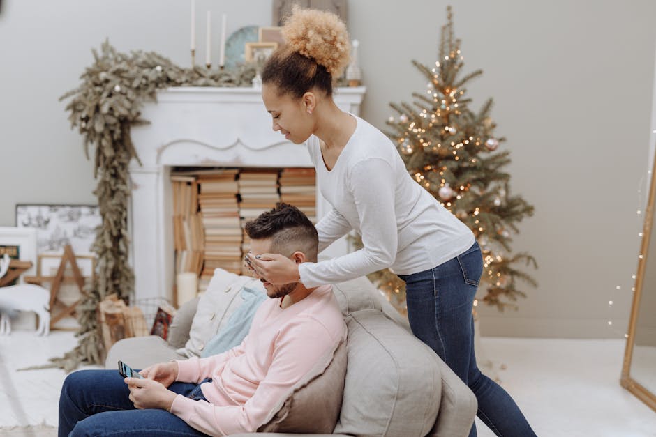A woman surprises a man in a cozy room decorated for the holidays by a Christmas tree.