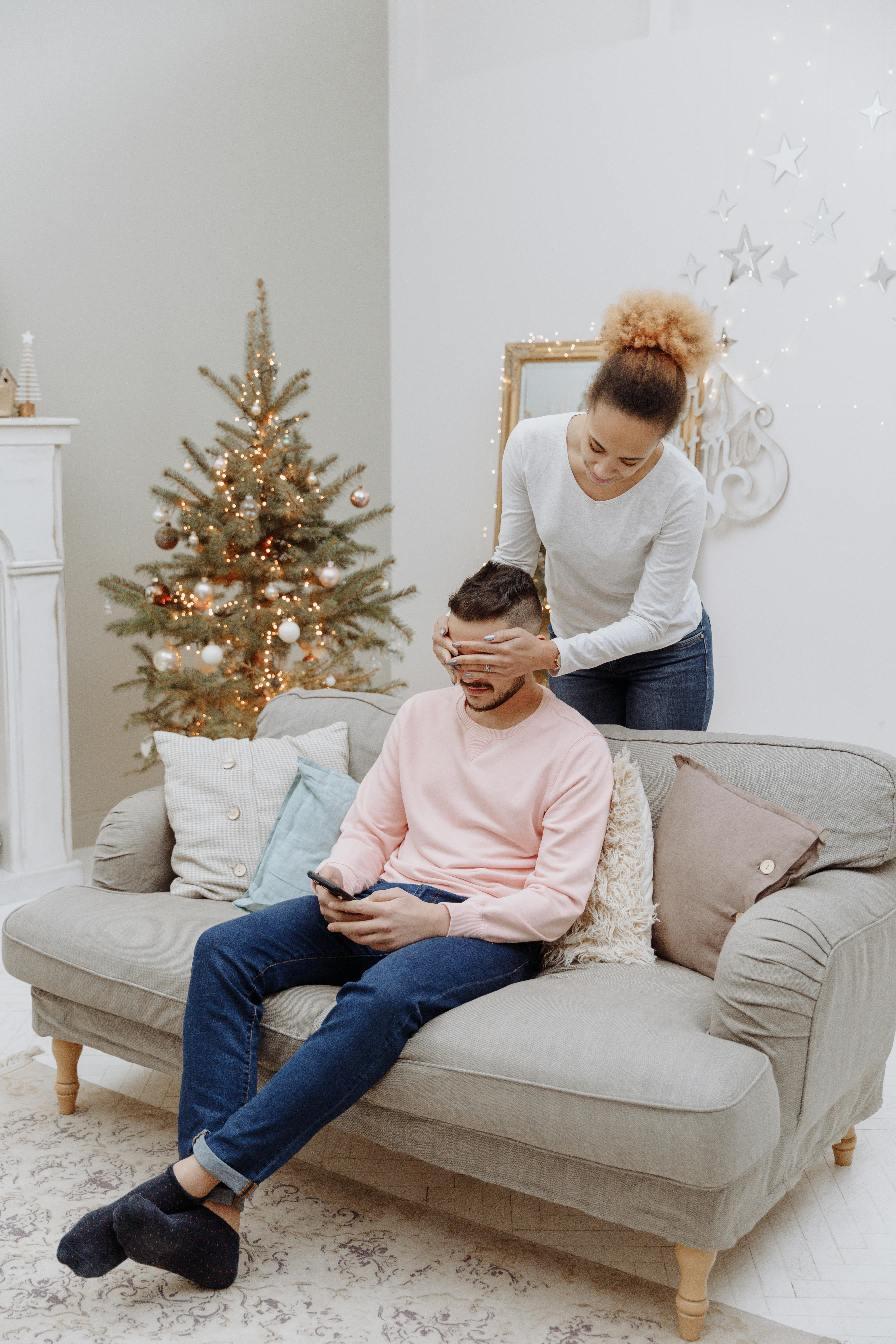 A woman playfully surprises a man during a cozy Christmas setting with a decorated tree.