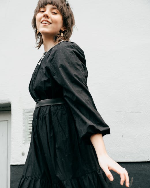 From below of young confident lady wearing black dress while dancing gracefully in street near building wall in daylight while looking at camera