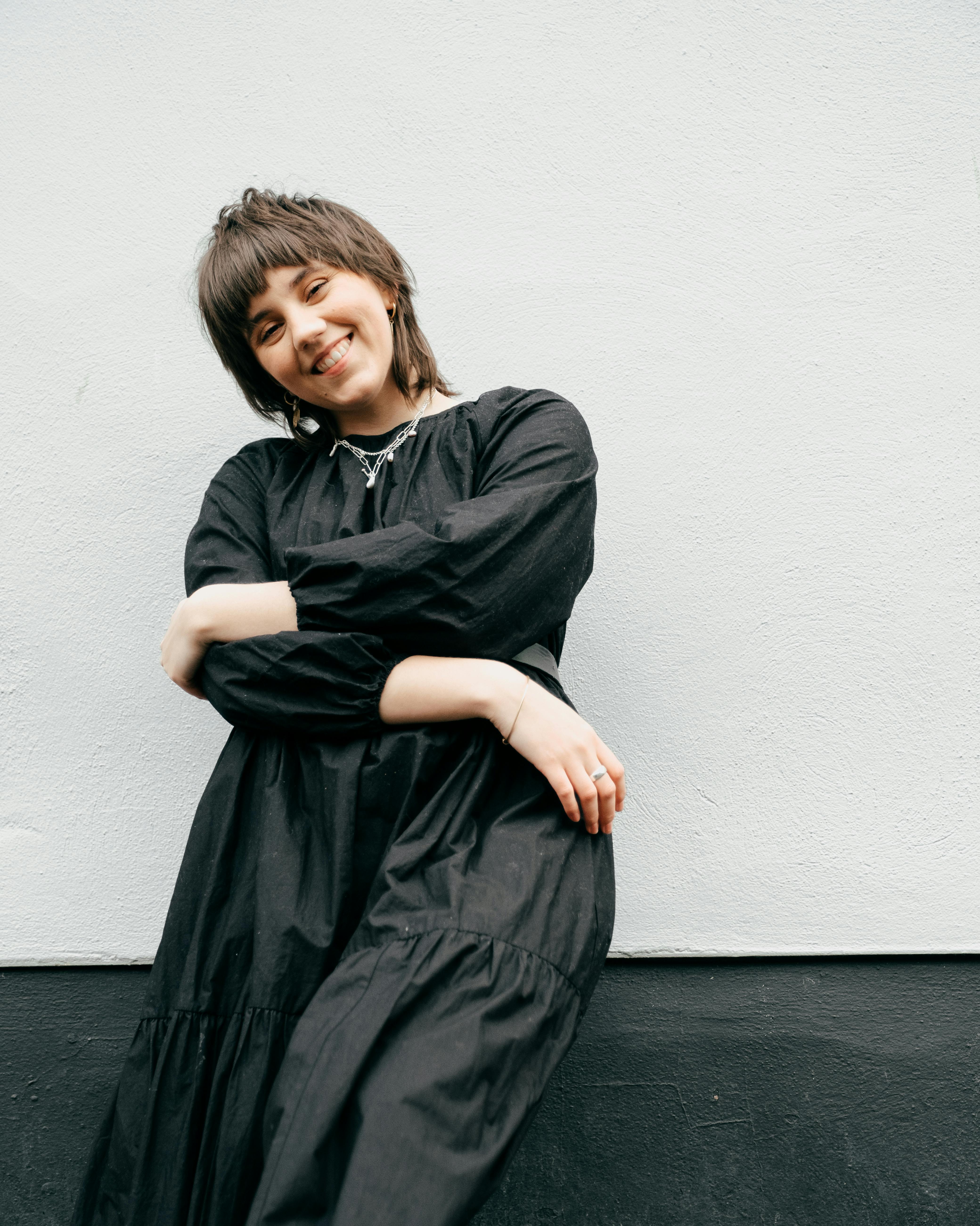 Happy young woman in black dress and accessories leaning on building wall in street while looking at camera with crossed arms in daytime