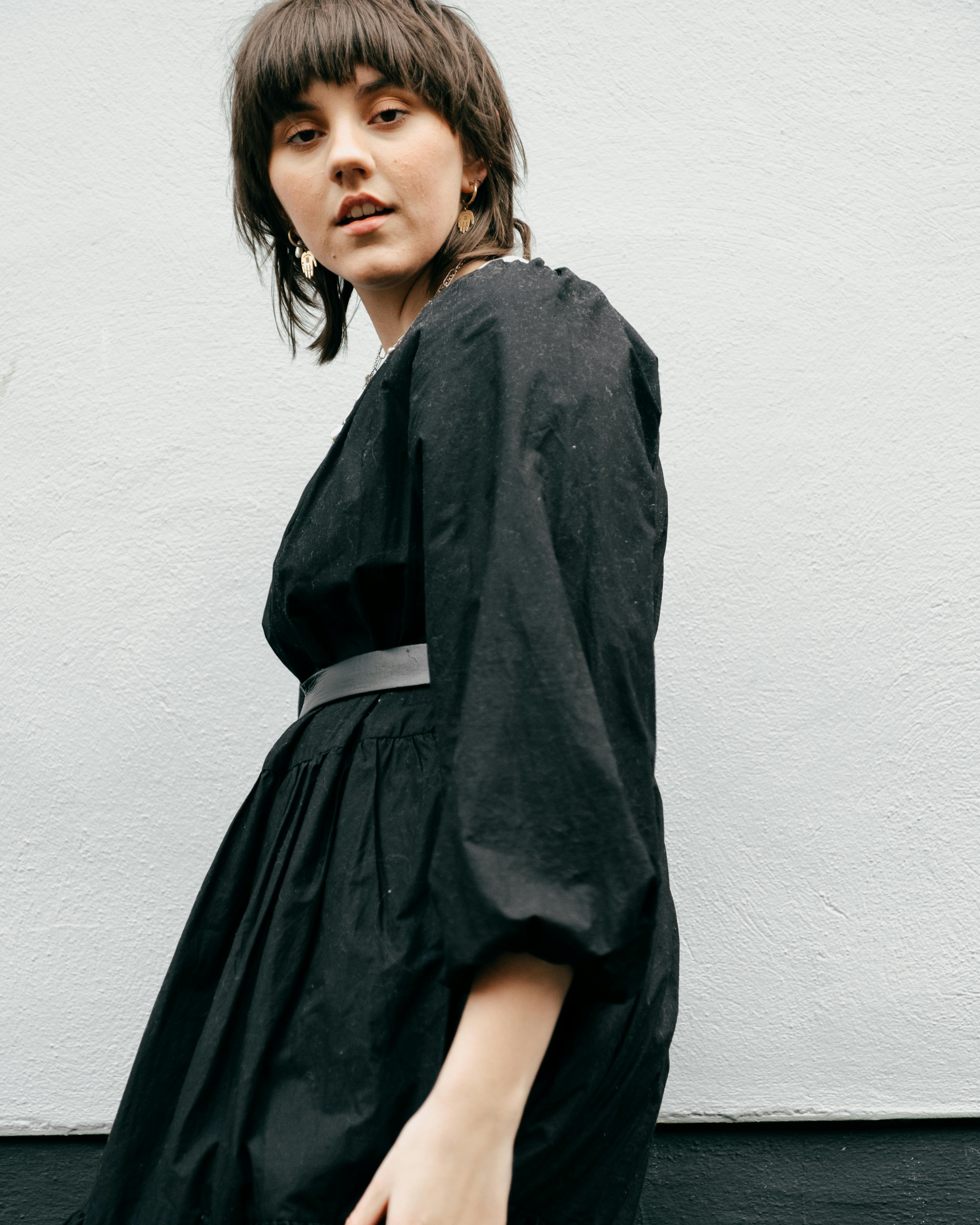 From below side view of young confident lady wearing black dress while standing in street near building wall in daylight and looking at camera