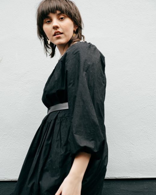 From below side view of young confident lady wearing black dress while standing in street near building wall in daylight and looking at camera