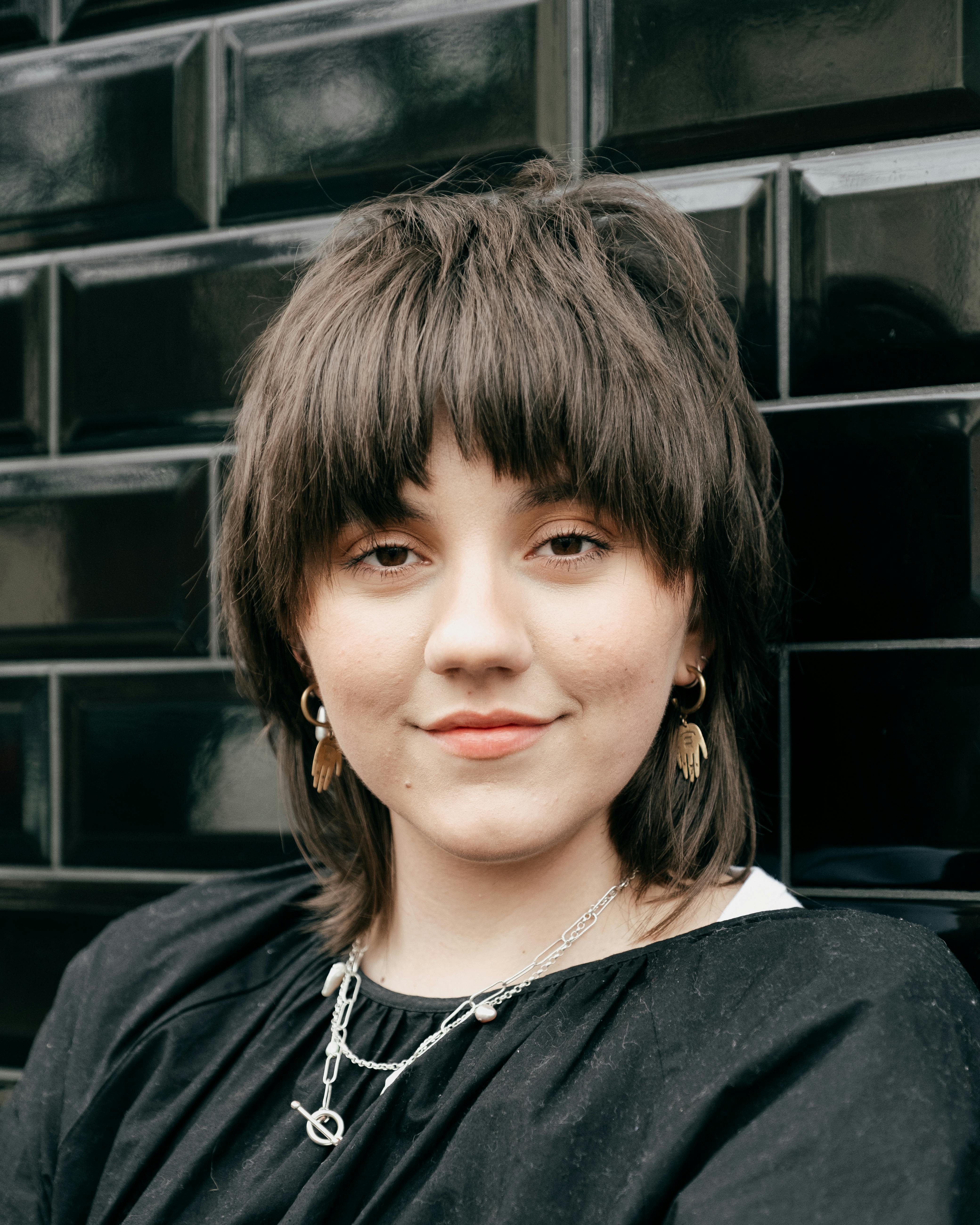 Calm young woman with short brown hair in casual clothes and accessories looking at camera against black brick wall in daylight