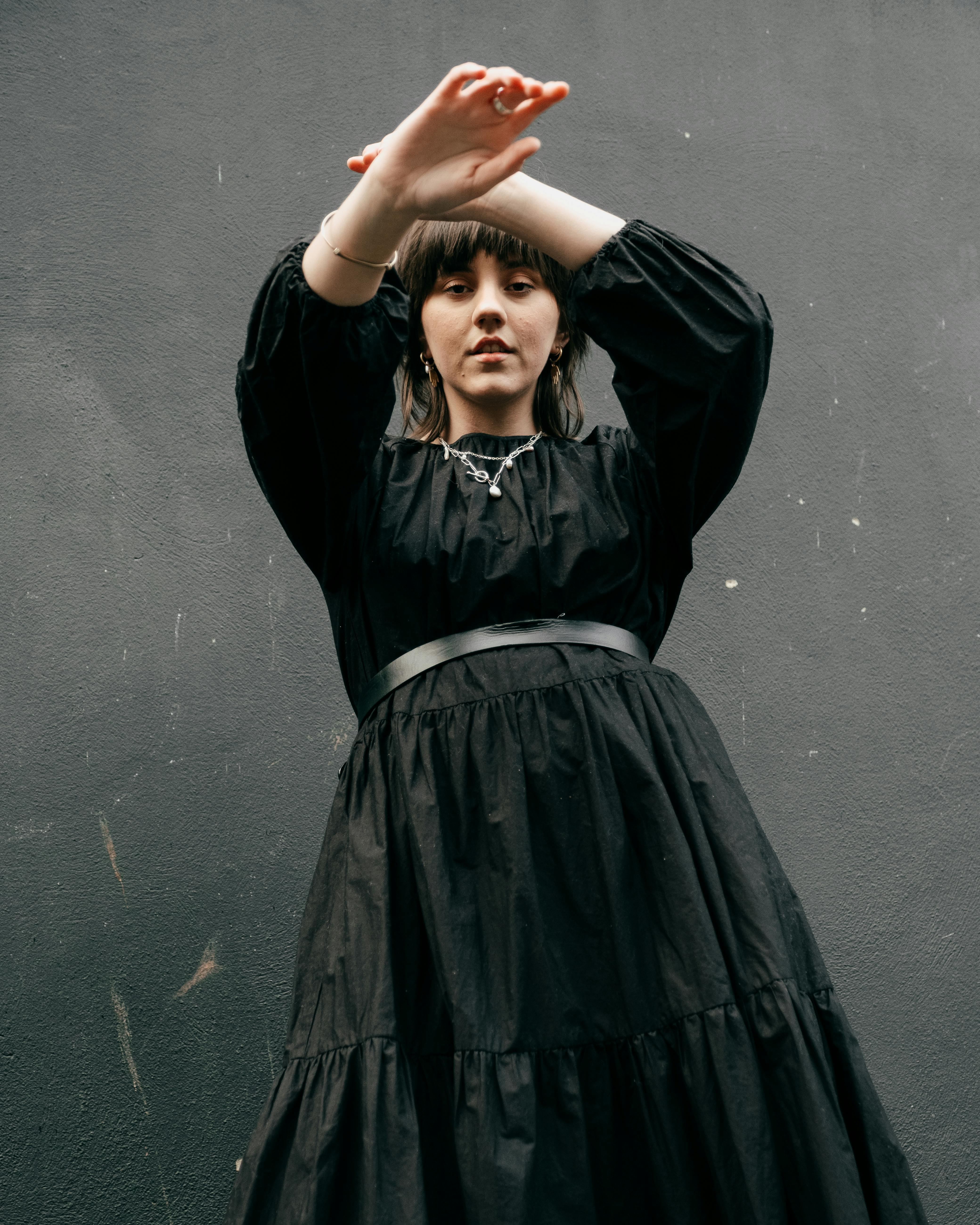 From below of pensive young female with brown hairs in trendy black dress standing with raised arms against black wall and looking at camera