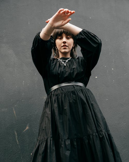 Calm female in black dress standing with raised arms against black wall