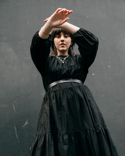 From below of pensive young female with brown hairs in trendy black dress standing with raised arms against black wall and looking at camera