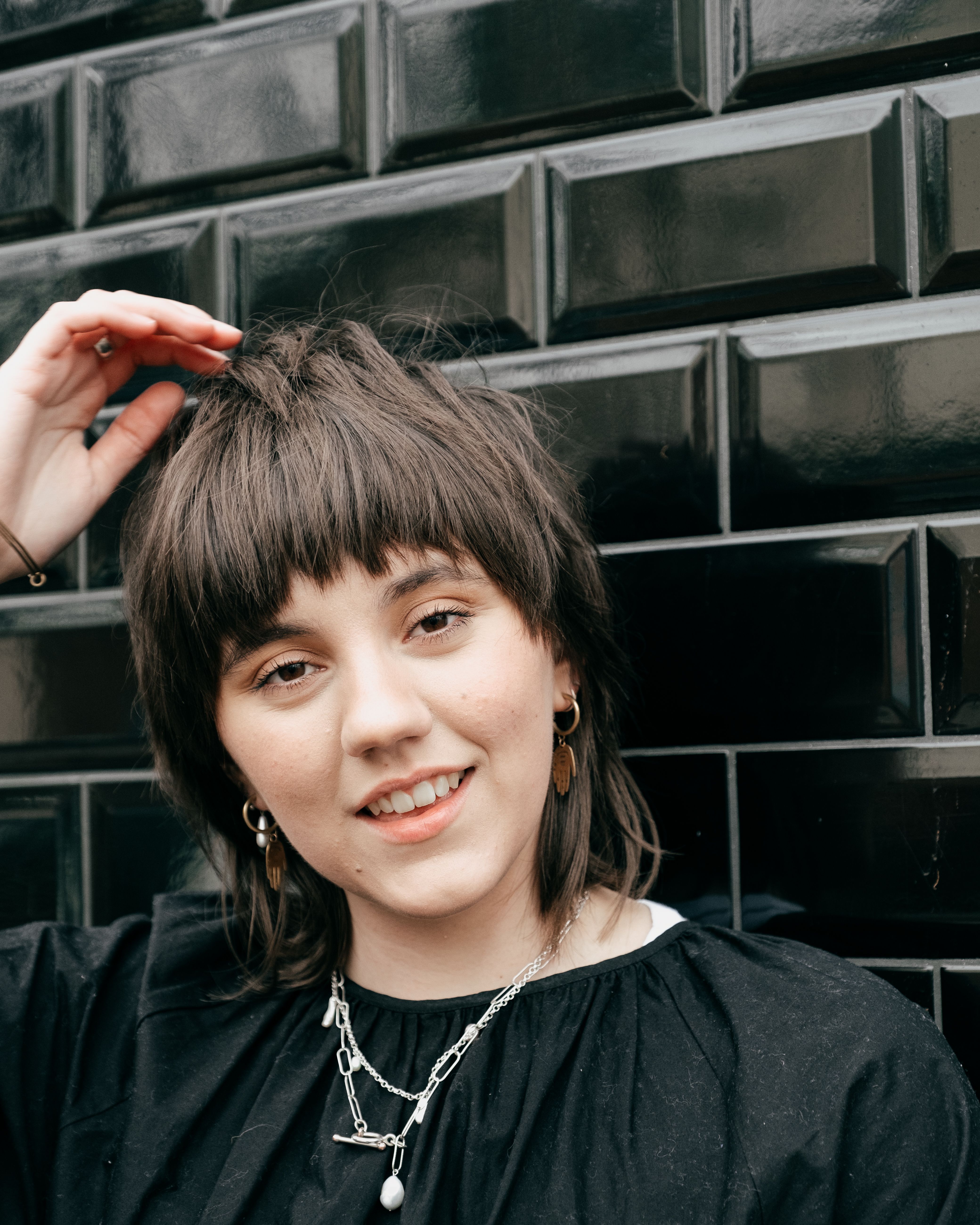 Cheerful young female with hairstyle in casual clothes touching hair and looking at camera against black brick wall in daylight