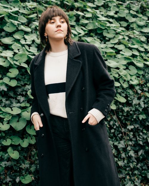 Free Serious female with hairstyle in stylish black coat standing with hands in pockets against green bush and looking away in daylight Stock Photo