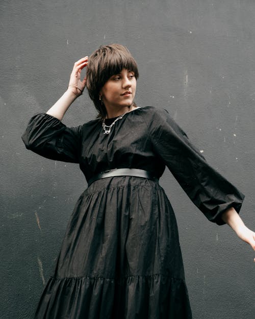 Charming young female with short dark hair in trendy black dress touching hair and standing against black wall in daylight