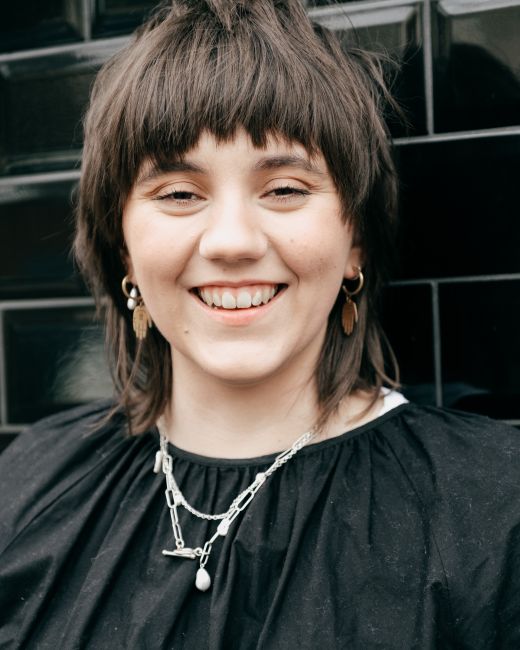 Cheerful young female with short dark hair in casual clothes and accessories smiling and looking at camera against black brick wall in daylight