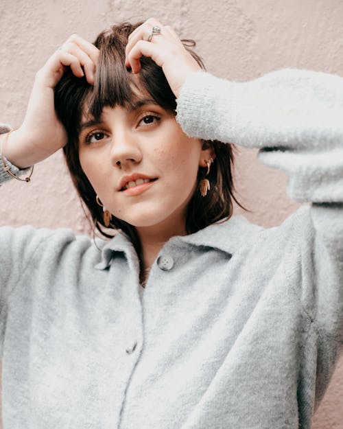 Charming female with brown eyes in casual clothes touching hair and looking away while standing near light concrete wall in daylight