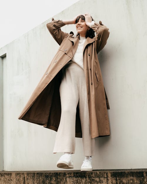 Low angle of full body of smiling female in trendy trench coat standing near building with light concrete wall and touching hair in daylight