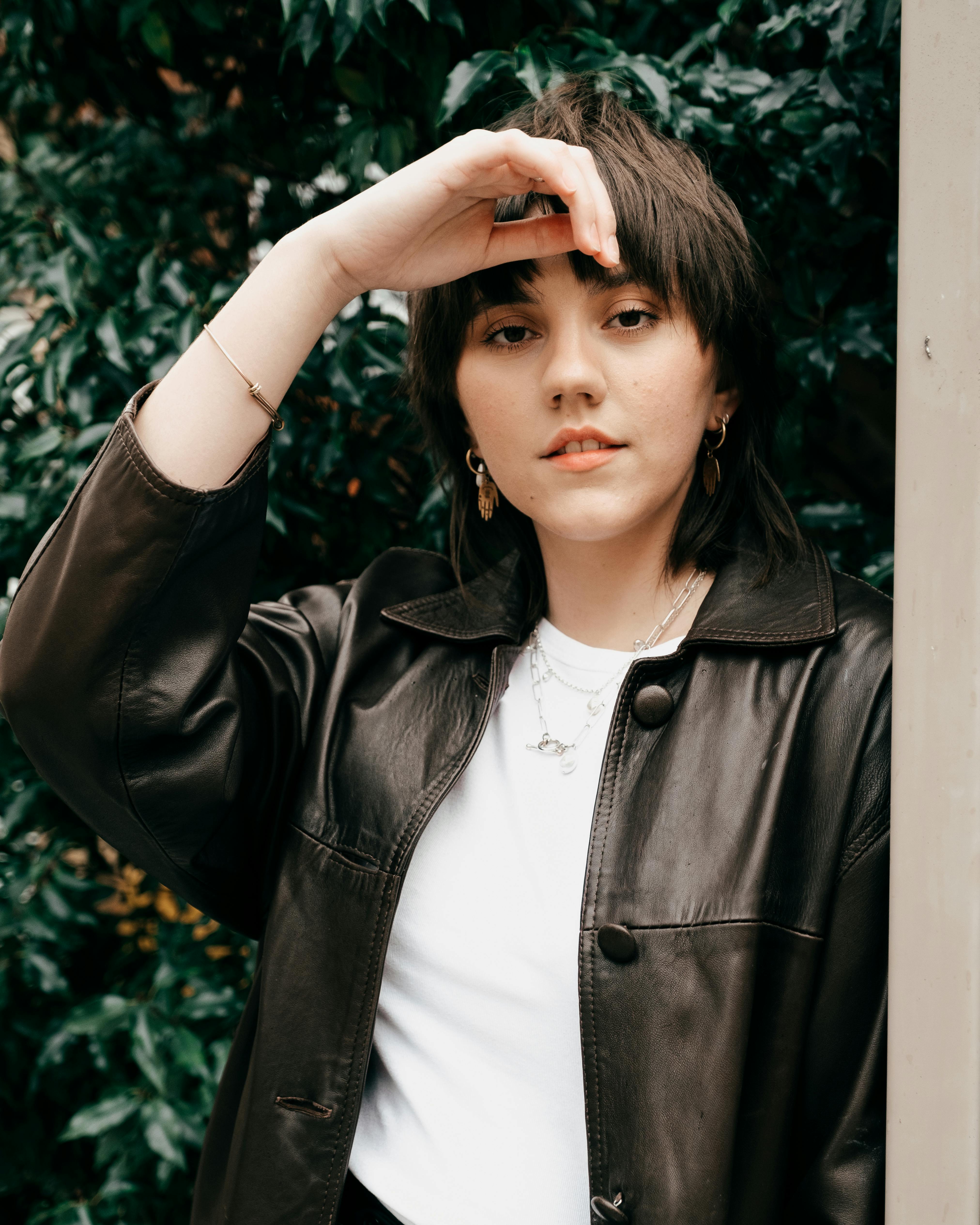 Stylish young female in black leather jacket standing with raised arm and touching head against green plant in daylight