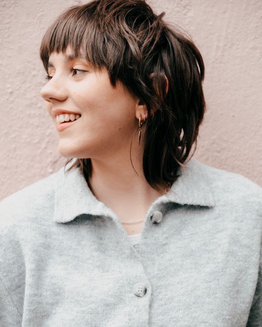 Positive young female with short brown hair in casual clothes smiling and looking away against light background in daylight