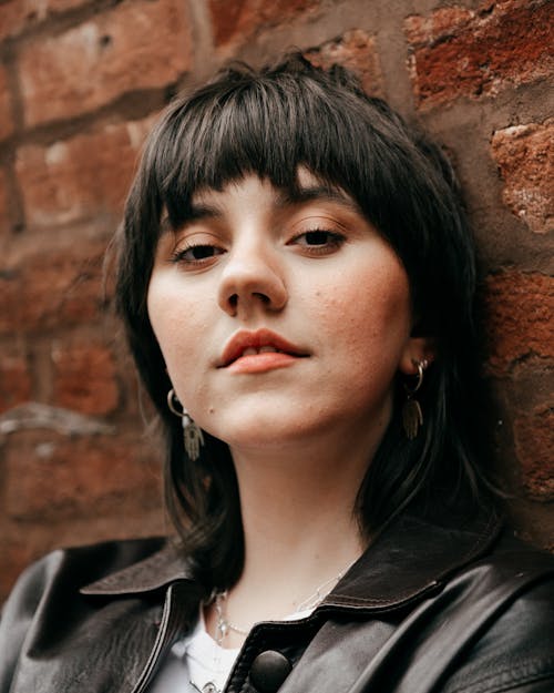 Calm young female with short dark hair in trendy clothes leaning on brick wall and thoughtfully looking at camera