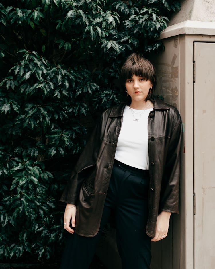 Serious Female Leaning On Wall Against Green Foliage Of Tree In Daylight